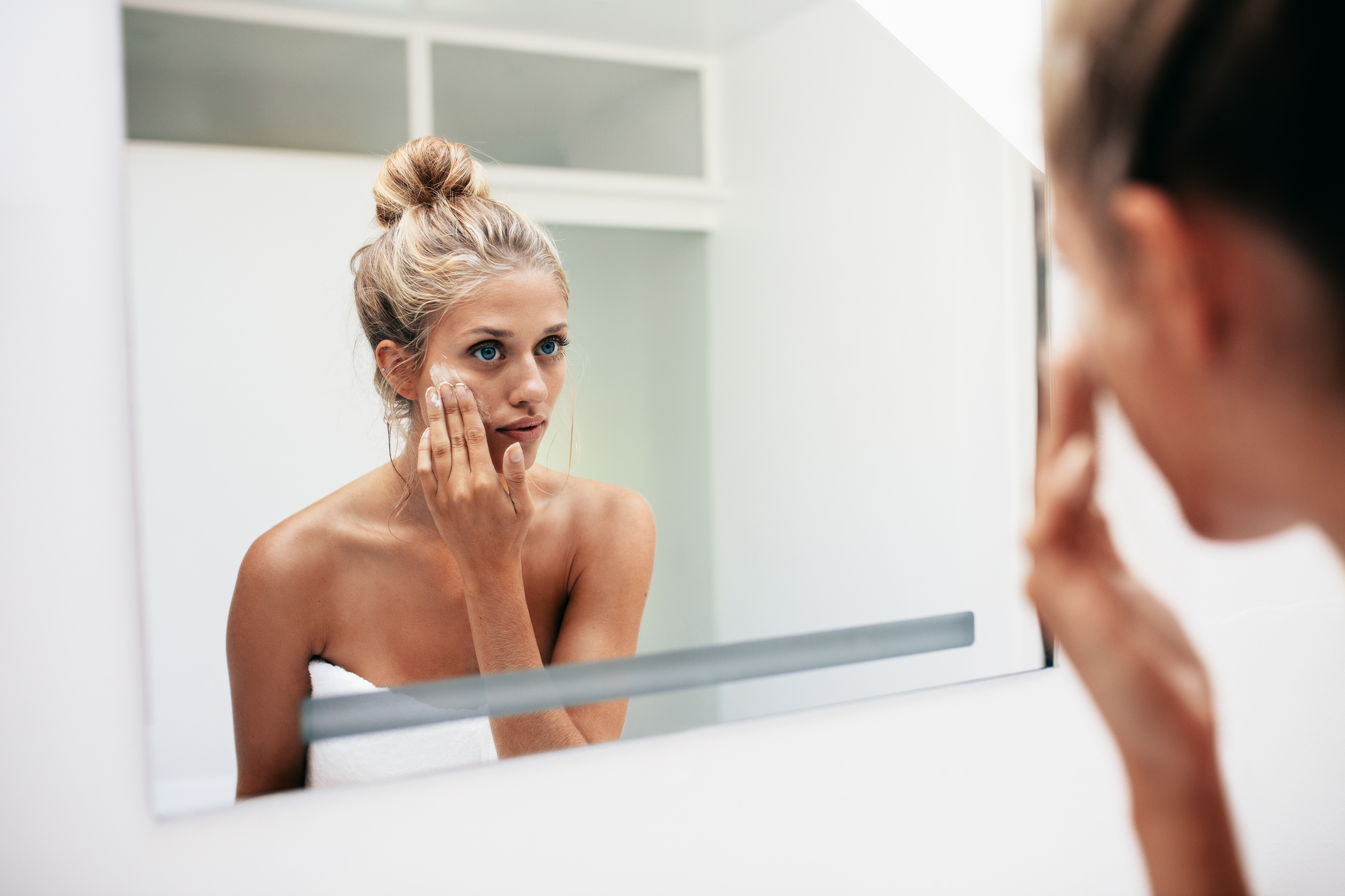 Reflection of a female in mirror rubbing cosmetic cream on her face. Female putting on moisturizer on her facial skin in bathroom.