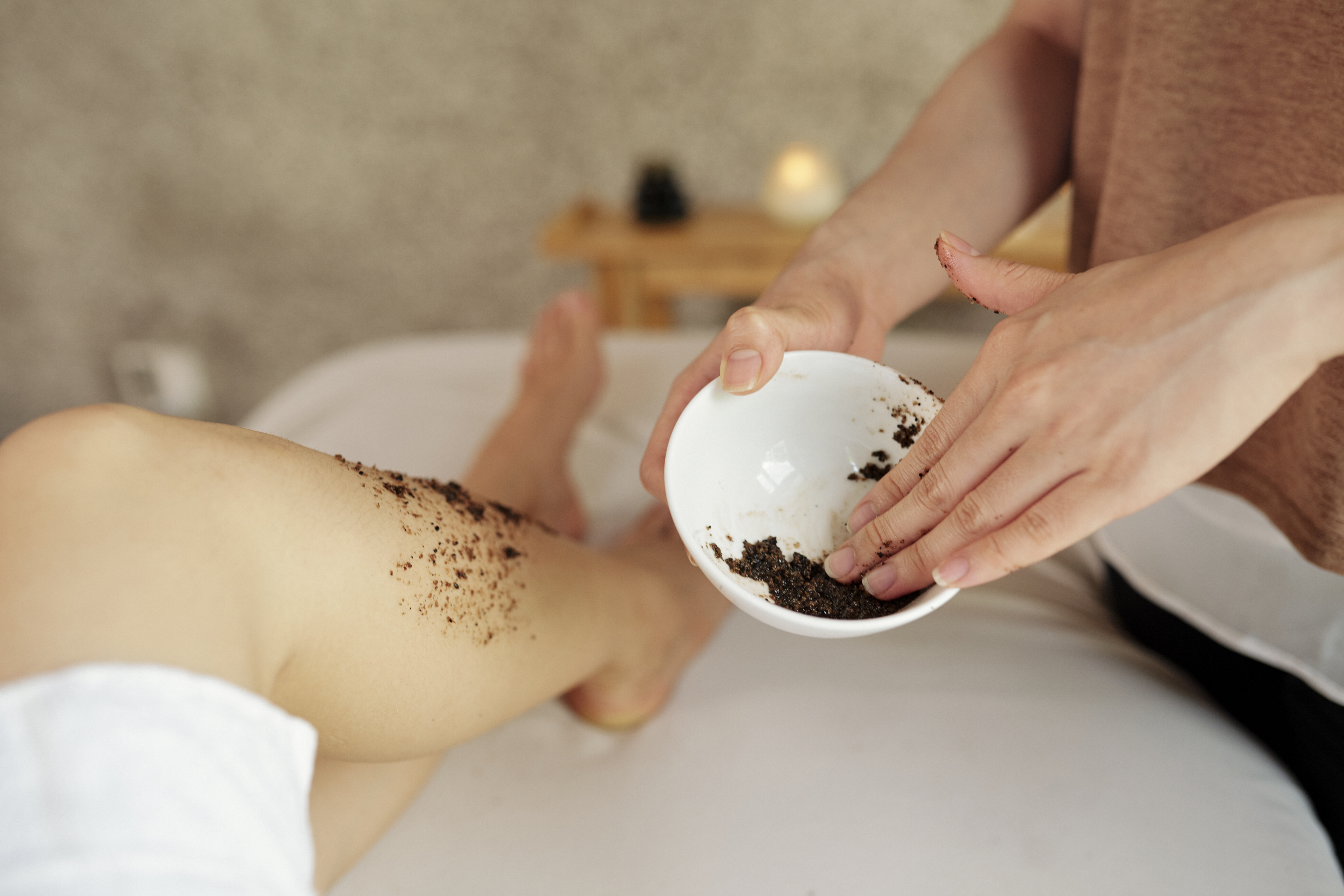 Close-up image of cosmetologist applying natural apricot scrub on legs of young woman relaxing in spa salon