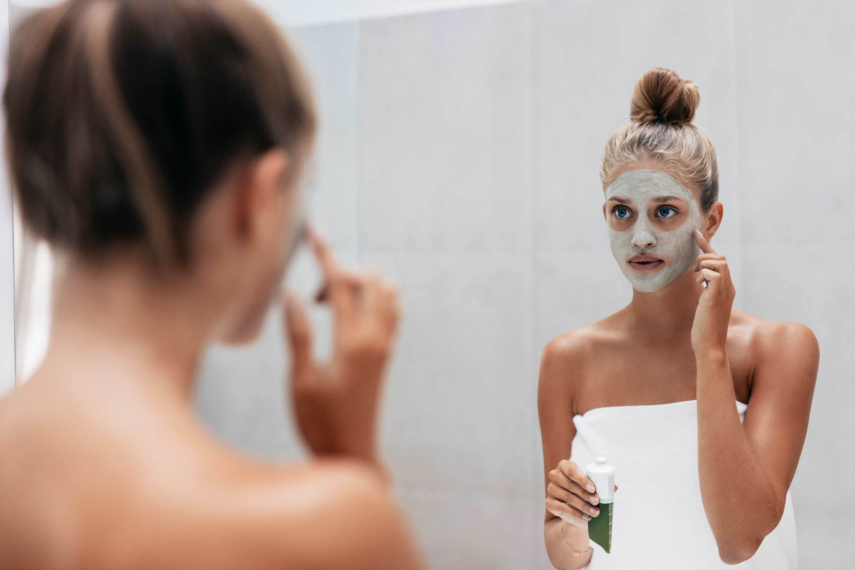 Beautiful young woman in bathroom applying mask on face looking at mirror. Girl taking care of her skin. Beauty treatment in bathroom.