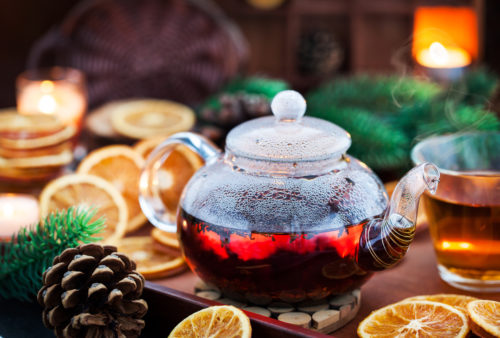 Glass teapot of hot black tea on cozy background with dried oranges and candles