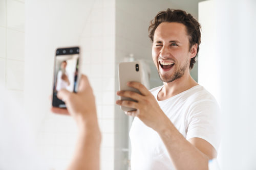 Attractive happy confident young man standing in front of the bathroom mirror, taking a selfie